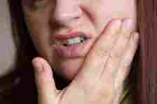 A person with long hair is holding their cheek, appearing to be in pain, possibly due to a toothache