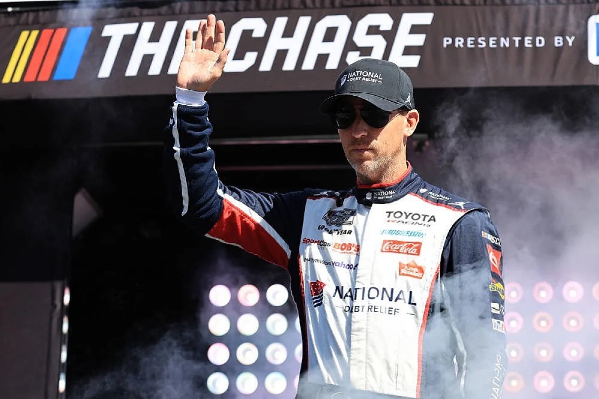 Driver in a navy and red Toyota Racing suit with National Debt Relief sponsor logos, waving at The Chase NASCAR event, wearing a black cap and sunglasses.