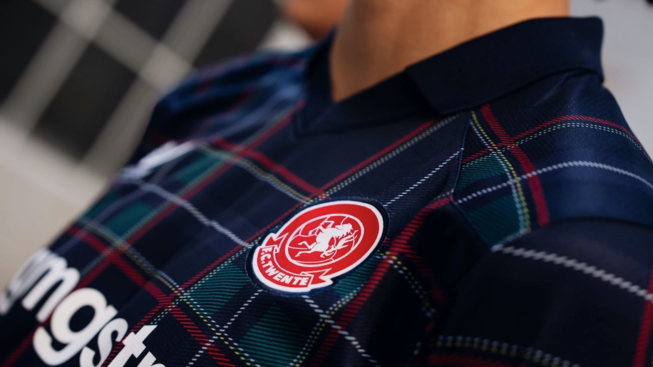 A tartan football shirt with the badge of Dutch club FC Twente on the upper right chest. The shirt is mainly dark blue and has threads of white, red and green running through it. The badge is mainly red, with a white horse in the middle and the name of the club in white text at the bottom.