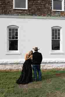 A woman in a black ruffled dress and a man wearing a cowboy hat and dark clothing stand closely together on a grassy area in front of a white brick building with two windows. The woman grasps the man's arm and leans her head towards him.