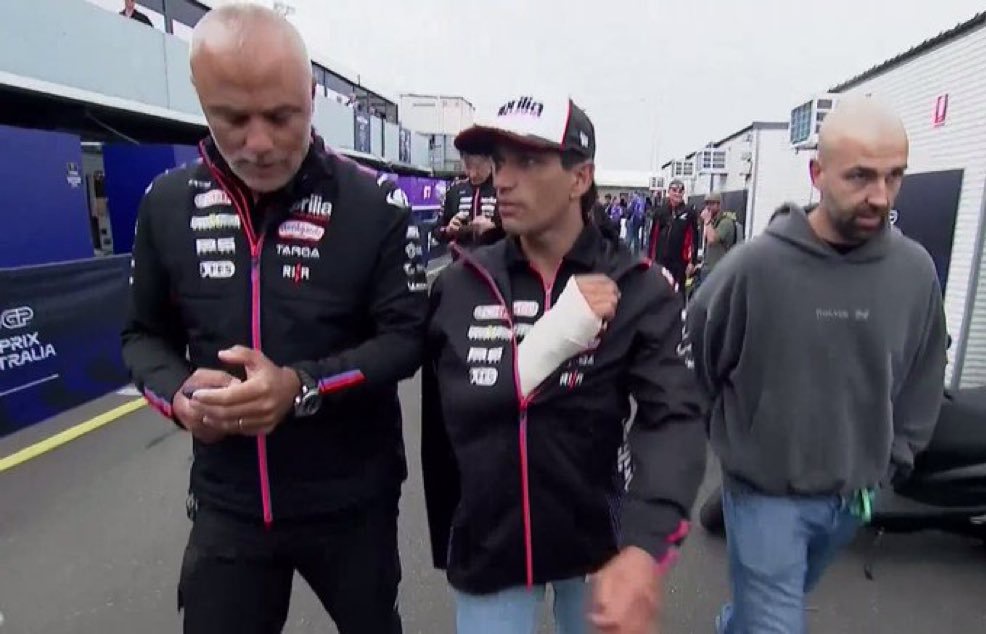 A man with a white cast on his arm wearing a black Aprilia team jacket and cap walks through the Australian GP paddock with two men beside him