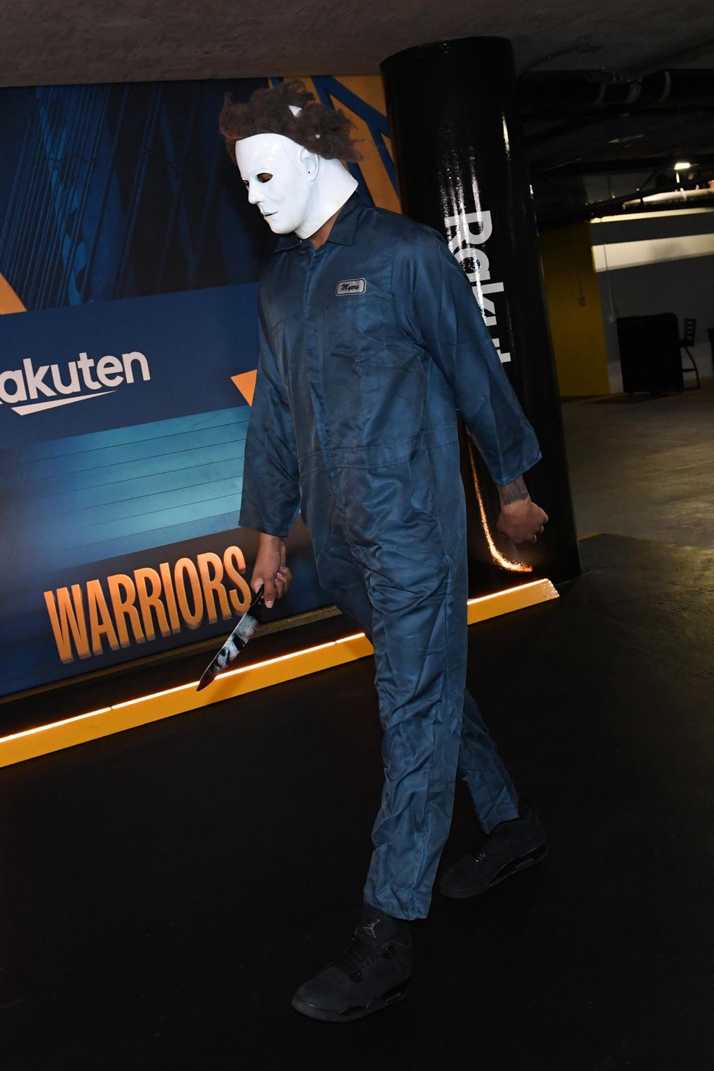 A person dressed as Michael Myers with a white mask, blue jumpsuit, and knife walks through a tunnel near a Warriors banner.