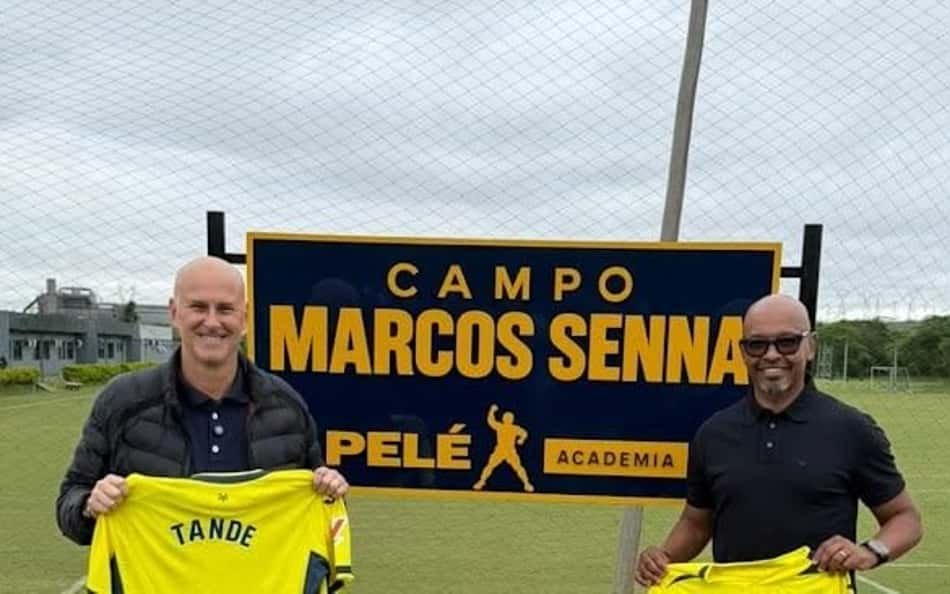 Duas pessoas seguram camisas amarelas diante de placa: Campo Marcos Senna Pelé Academia, em campo de futebol.