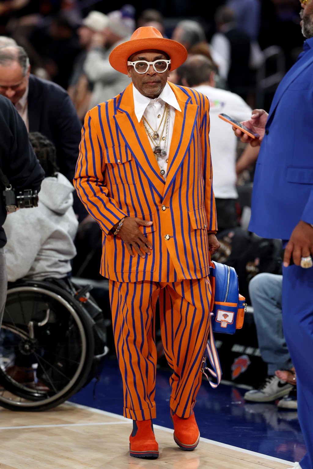 Spike Lee stands on the basketball court wearing a bright orange and blue striped suit, matching hat, and thick white glasses.
