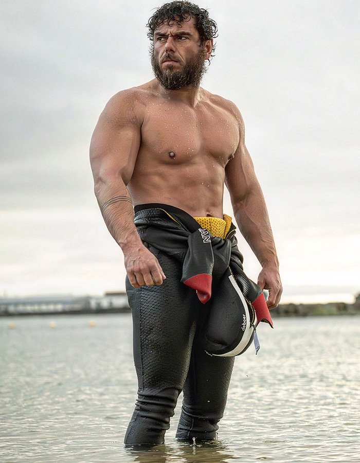 Extreme swimmer standing in shallow ocean water wearing a partially unzipped wetsuit.