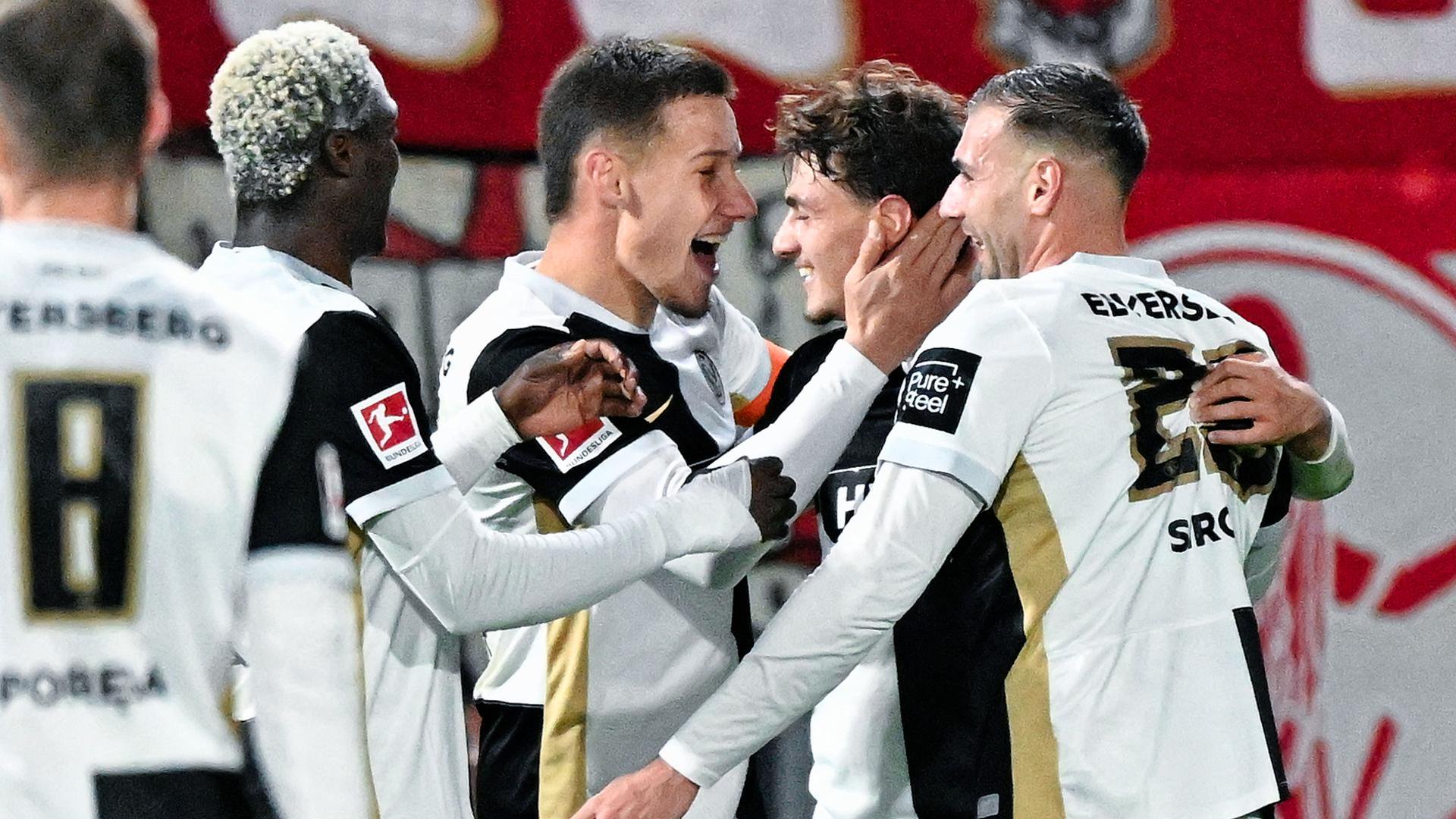 Das Bild zeigt mehrere Spieler des SV Elversberg in der Ursapharm-Arena an der Kaiserlinde. Sie jubeln über das Tor zum 3:0 und fallen sich in die Arme. Im Hintergrund ist ein Werbebanner zu erkennen.