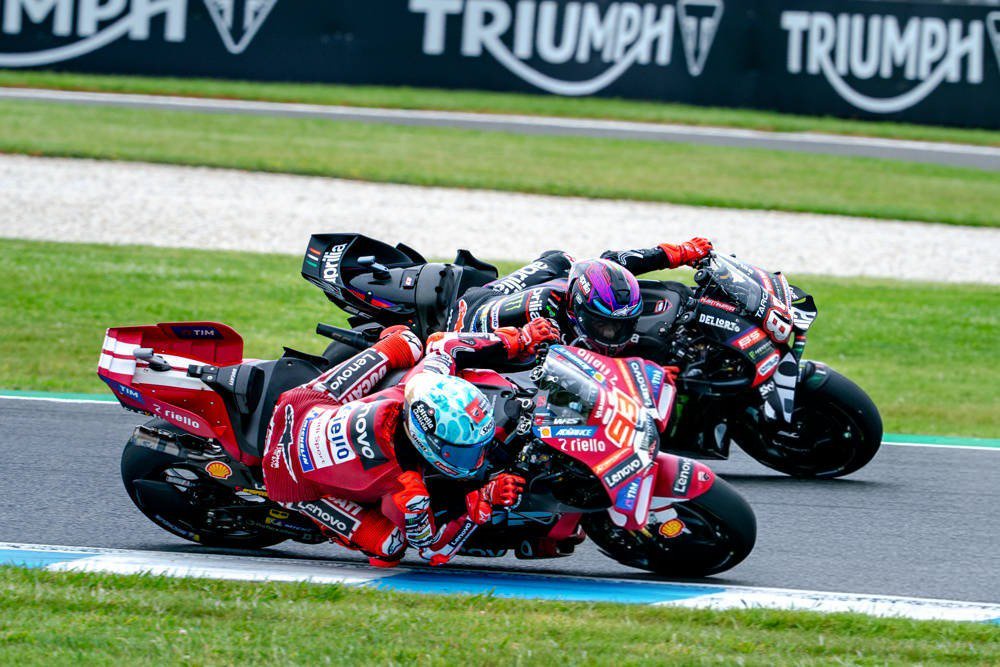 Ducati and Aprilia MotoGP riders racing closely on a track, one in red Ducati leathers with Lenovo branding, another in dark Aprilia livery behind