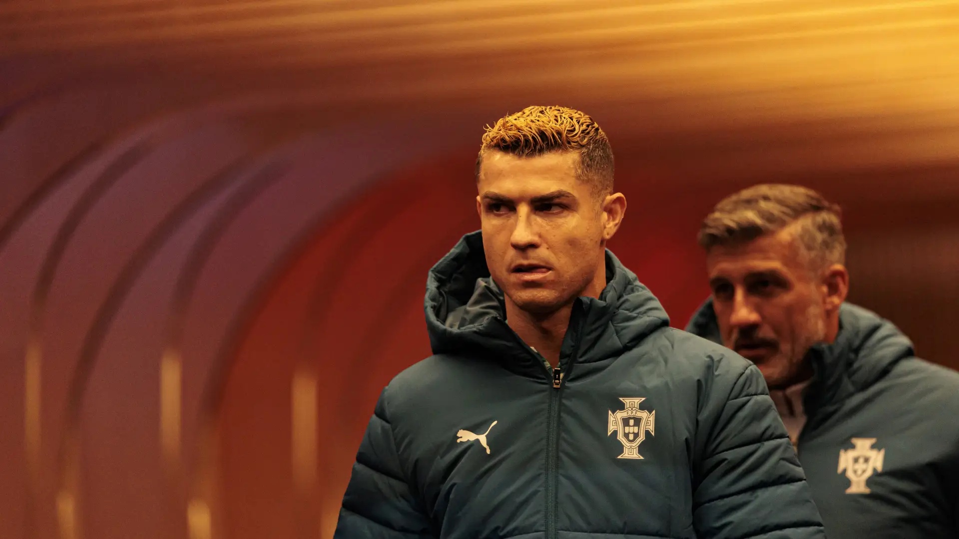 Cristiano Ronaldo, com cabelo loiro e jaqueta azul-marinho da seleção portuguesa, caminha com expressão séria. Um homem de barba grisalha o segue, parcialmente visível, em um túnel iluminado por tons de laranja e vermelho