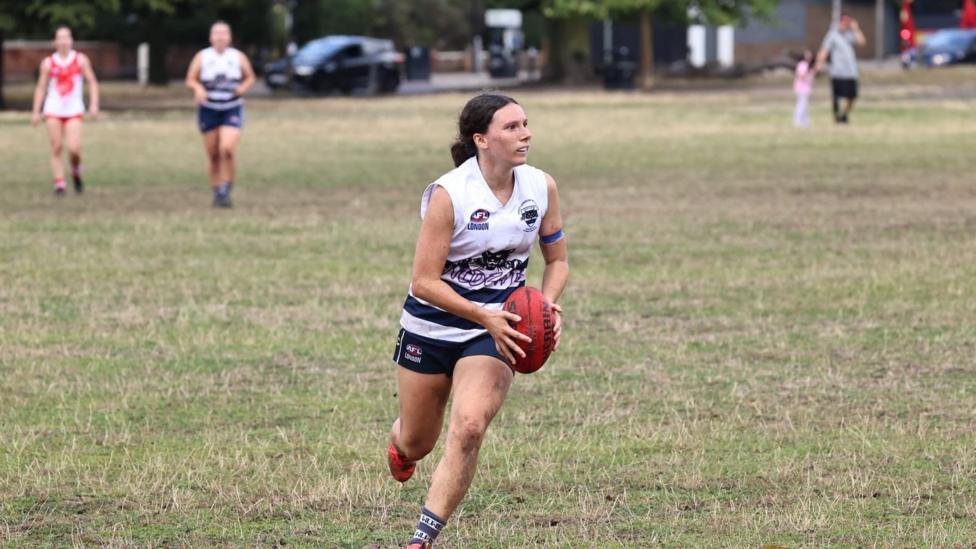 A player in a white-and-navy sleeveless sports kit runs across a grass field holding a red Australian rules football. Two other players are visible in the background, with trees, cars and pedestrians beyond the pitch.