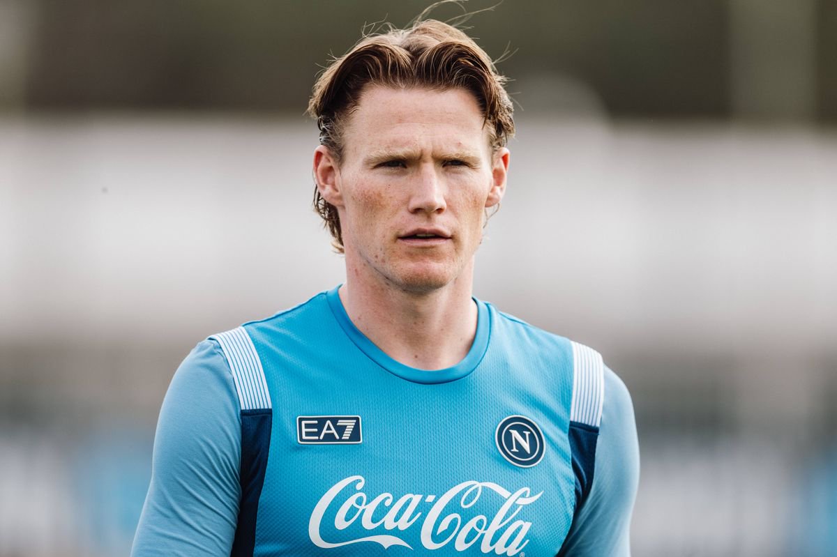 An individual, clad in a blue sports jersey with a prominent Coca-Cola logo, is seen standing on a field, with a serious expression and a blurred background.