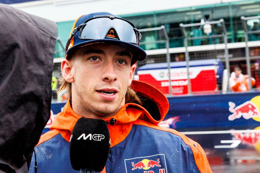 A rider in a Red Bull KTM jacket and cap speaks into an MCP microphone in the paddock, with a wet track behind him.
