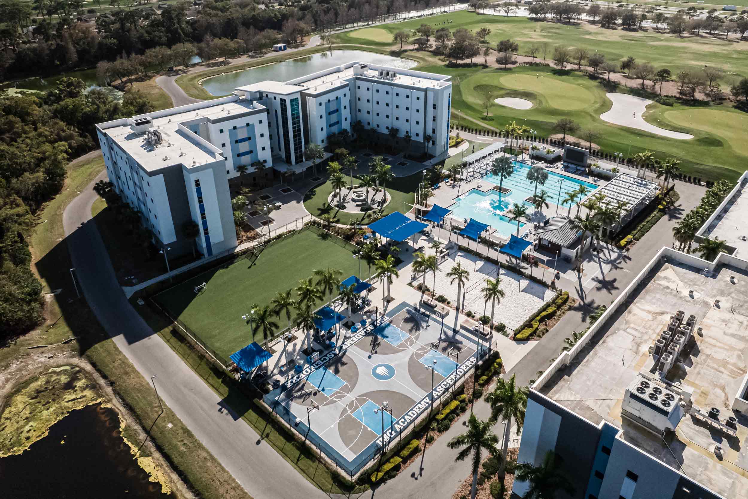 IMG Academy's south dorm is flanked by a swimming pool, a green recreational area, a sand volleyball court, and a basketball court.