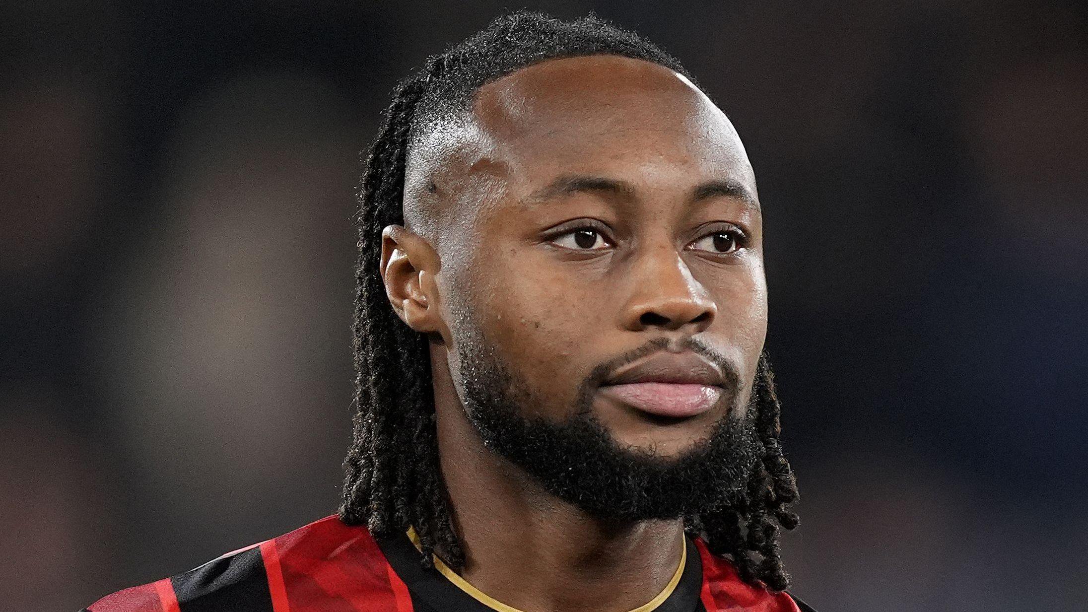 A close-up photograph of a Semenyo wearing Bournemouth's red-and-black striped shirt with gold trim around the collar. He has long black braids falling towards the back of his neck, with a beard and moustache.