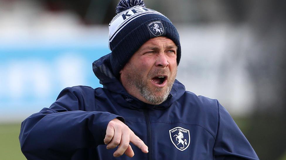 Matt Walker shouting and pointing instructions, wearing a blue and white Kent CC beanie hat and blue padded training jacket