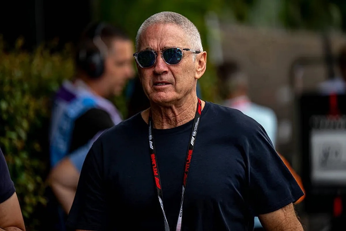 A man with short grey hair, dark sunglasses and a navy t-shirt, wearing a black and red lanyard, in a paddock area with blurred people behind him.