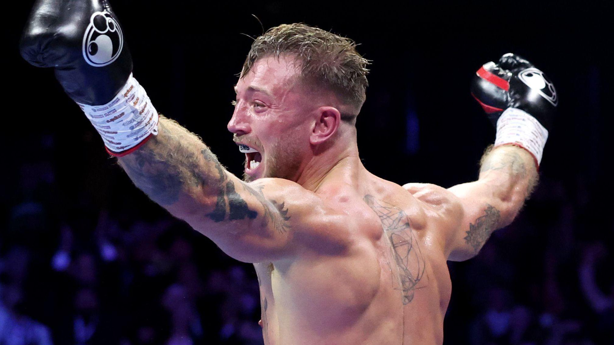 Brad Pauls stands with his back to the camera and his arms held in the air in celebration. He is wearing boxing gloves and his mouth is open