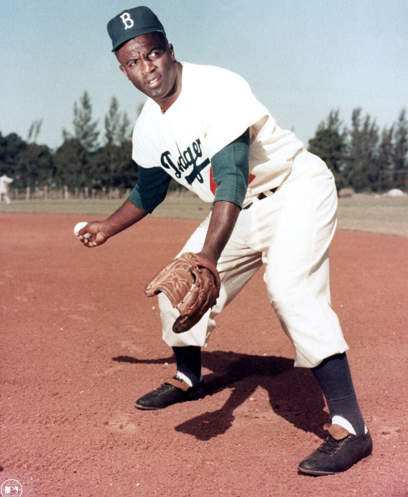 A baseball player wearing a vintage white uniform and blue cap is poised on a dirt field, glove on one hand, ball in the other. Trees and a clear sky fill the background.