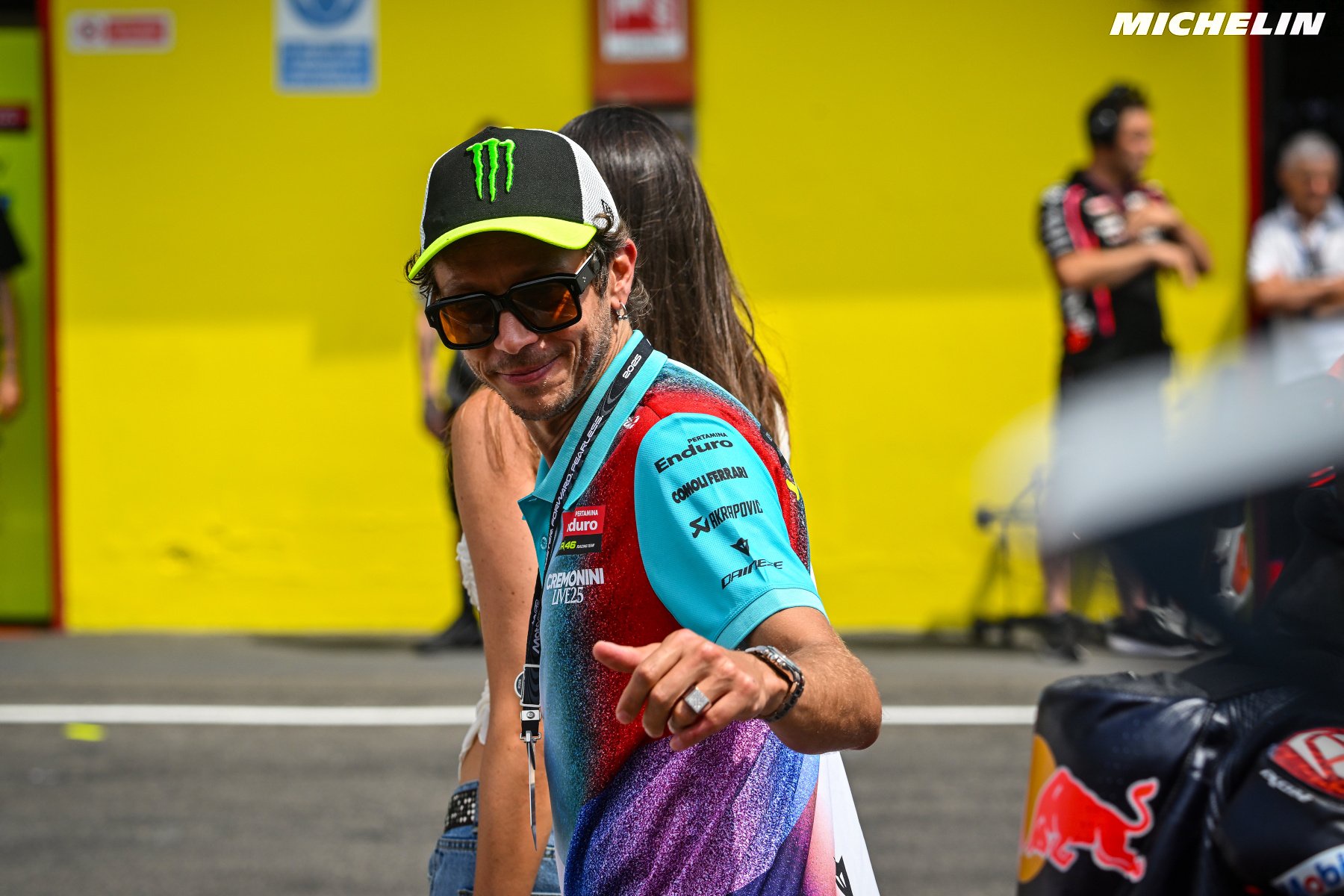 A smiling man in sunglasses and a monster-logo cap wears a teal Enduro team shirt in a MotoGP paddock, beside a Red Bull-liveried bike.