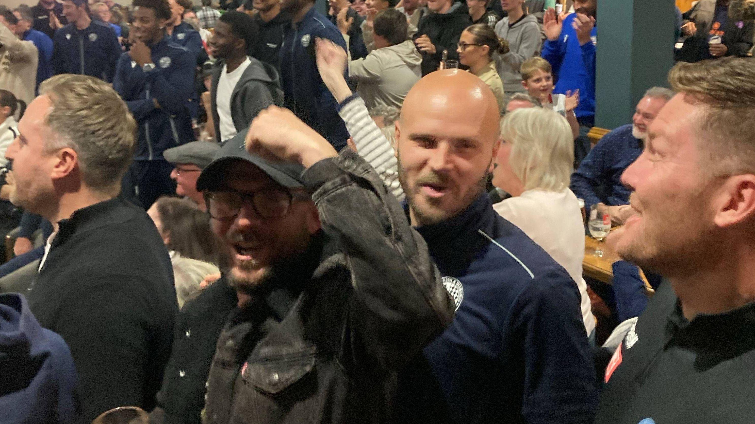 Rob Usher and Elliot Sandy celebrate inside Rushden & Diamonds' clubhouse. The men are surrounded by other men, women and children. Rob is on the left, with a dark beard and dark-framed glasses, wearing a black baseball hat and jacket and raising his left hand. Elliot is on the right, with a light brown beard and wearing a blue tracksuit. 