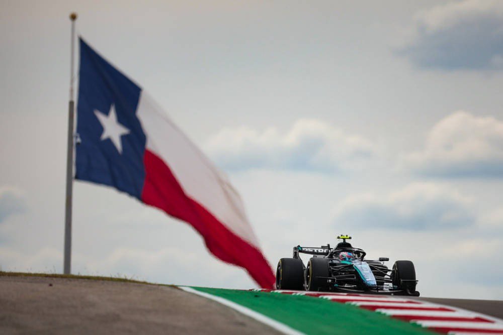 Formula 1 car in dark livery with yellow helmet stripe racing past a Texas flag at Austin's circuit, United States Grand Prix
