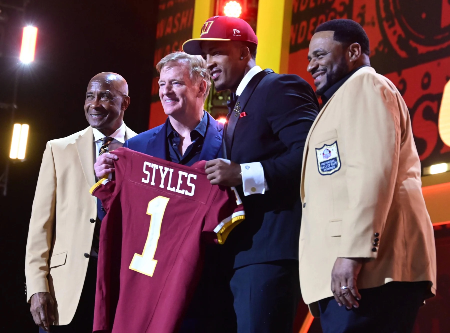 Four men stand onstage, one holding a maroon football jersey labeled “STYLES 1.” Two men wear tan jackets with Hall of Fame patches as the 2026 NFL Draft highlights the Rams' Big Ten prospects. Alt text generated by AI.