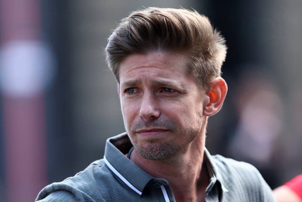 A man with swept-back brown hair and stubble wears a grey collared shirt, looking off to the side with a sombre expression in a paddock setting.