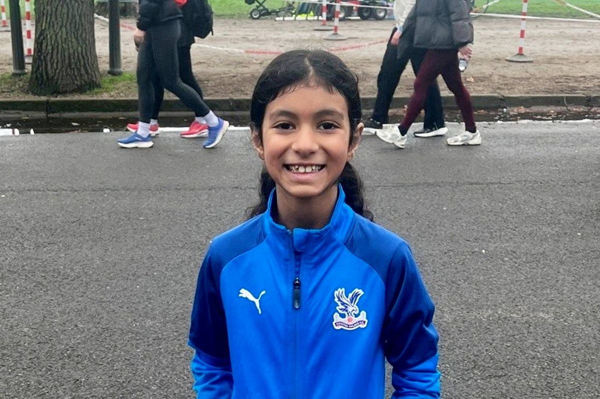 A young girl, clad in a blue jacket and white socks, stands confidently in an urban street, smiling broadly and gazing ahead. The background features a blurred image of pedestrians, suggesting a lively urban environment.