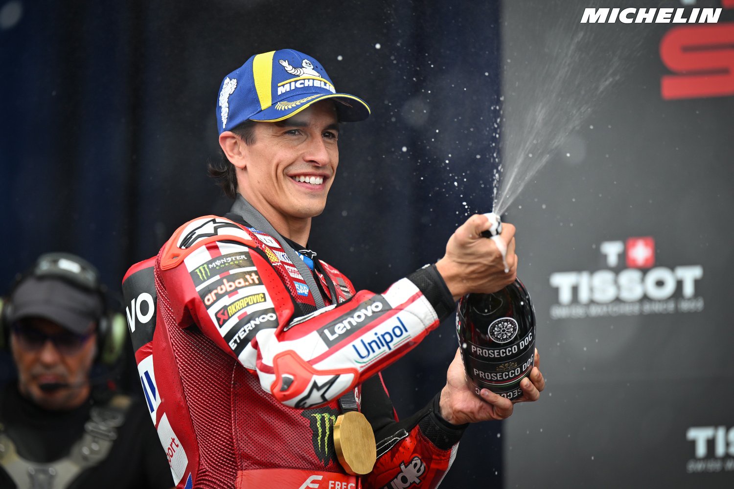 A rider in red Ducati leathers, Pramac-style sponsors visible, smiling and spraying Prosecco from a bottle on the podium, with a Michelin banner behind.