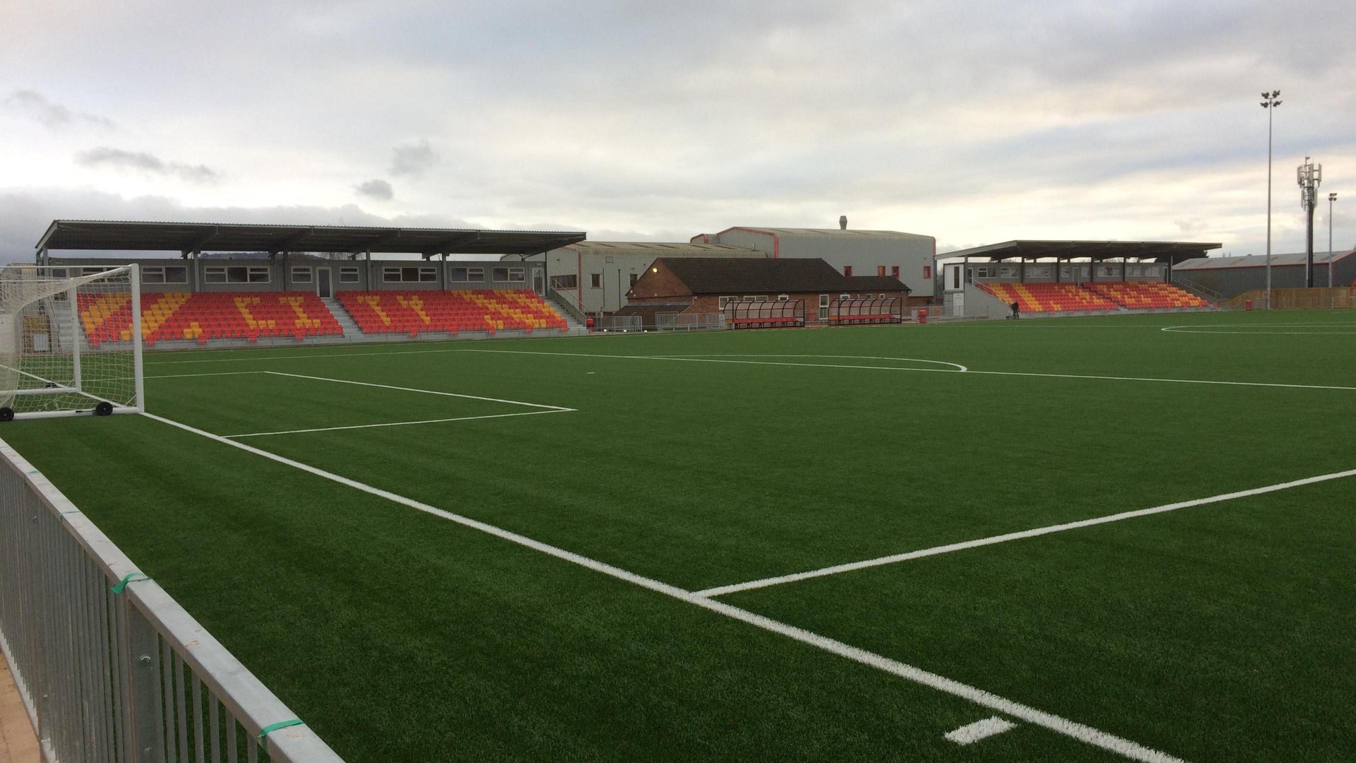 A non-league football ground, with the goal to the left of the photo and stands at thew back of the photo. The sky in the photo is grey and cloudy.