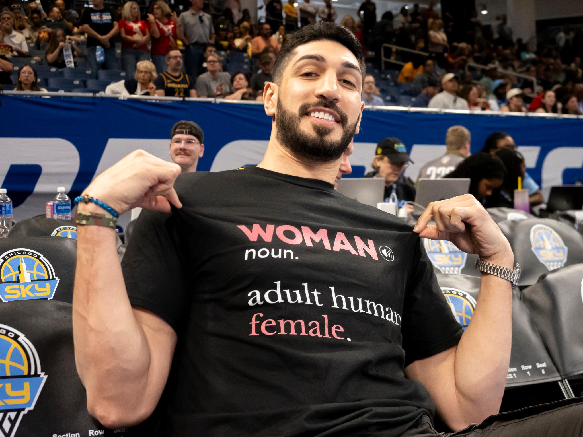 Enes Kanter Freedom sitting courtside wearing a t-shirt that reads "WOMAN noun. adult human female."