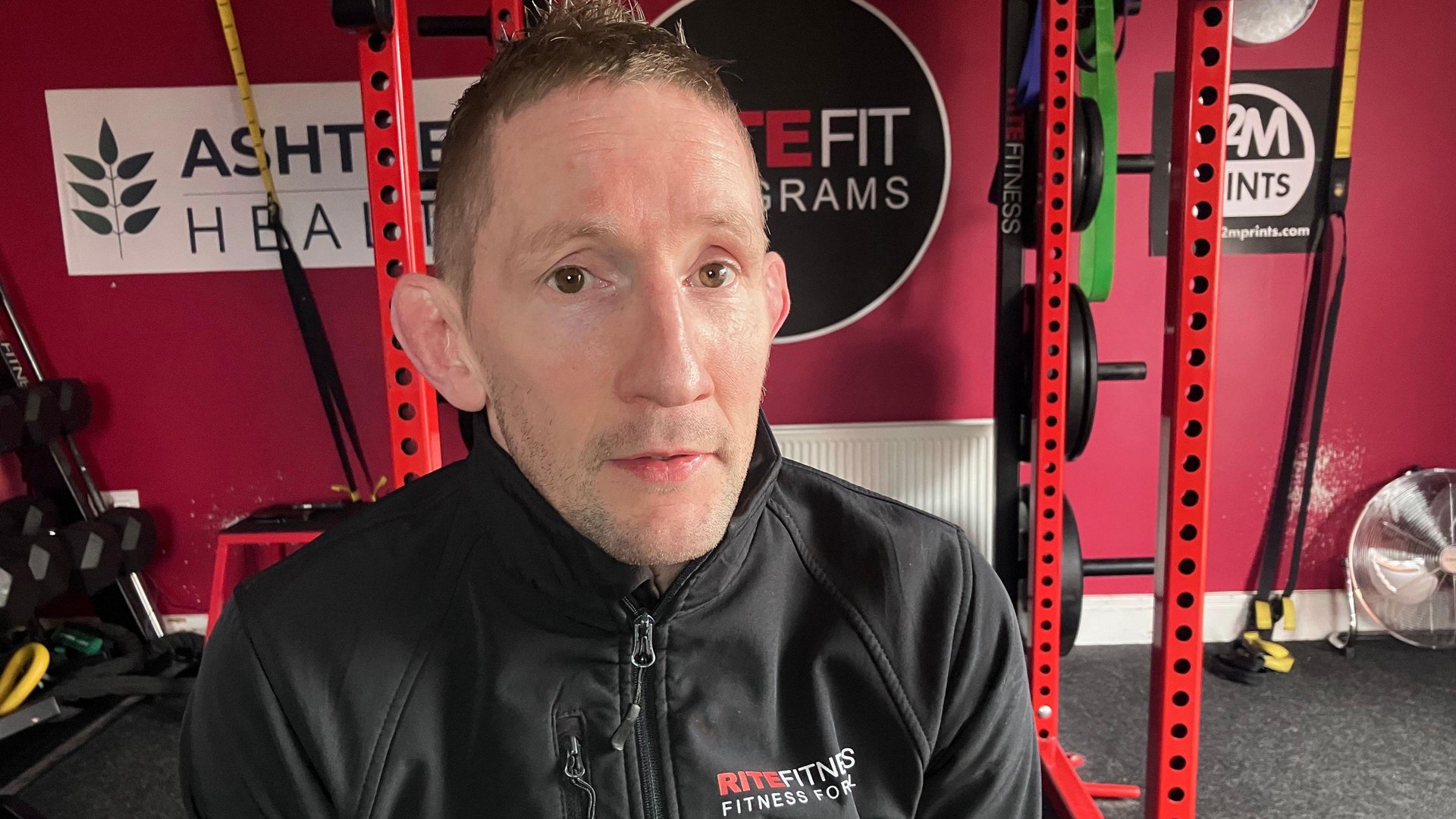 A man wearing a black sports jacket which says Rite Fitness while sitting on a piece of apparatus in a gym.
