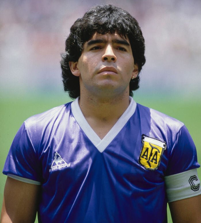 Diego Maradona wearing a blue jersey with the letters "AA" on the emblem stands on a field. He has curly dark hair and is looking ahead. The background is blurred with a crowd.