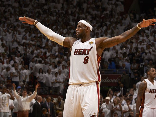 Basketball player in a white Miami Heat uniform, number 6, extends both arms wide. He's on a basketball court with a crowd wearing white in the background.