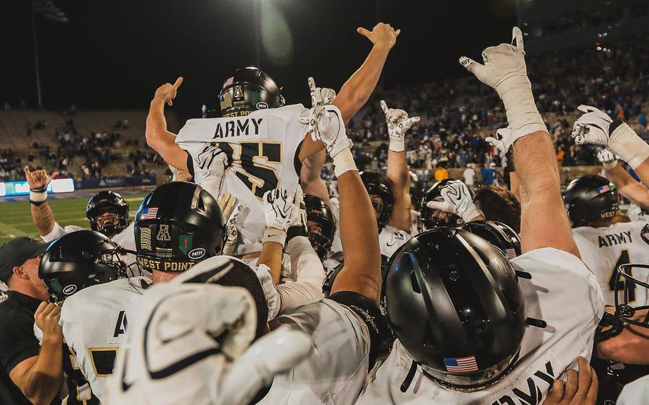 A college football player in a white jersey and black helmet, seen from behind, is lifted up and carried off the field by teammates.