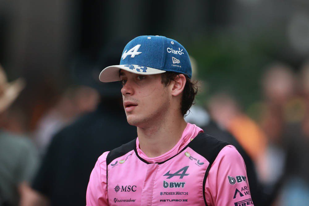 Alpine F1 driver in pink race suit and blue cap with BWT and MSC logos, standing in a paddock area.
