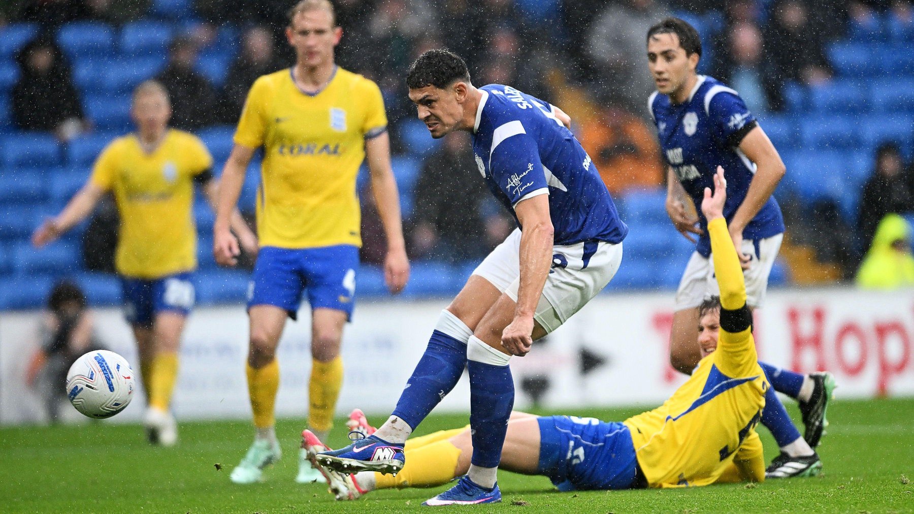 The ball has just been kicked as Yousef Salech in blue shirt and white shorts  scores for Cardiff surrounded by yellow and blue-clad defenders, one on the ground with arm in air
