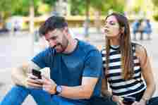 Woman looking over man's shoulder as he reads his smartphone