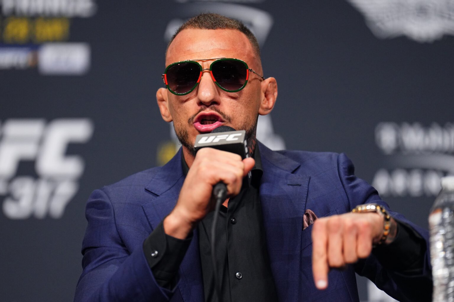 Renato Moicano in a navy suit speaks into a UFC microphone at the UFC 317 press conference