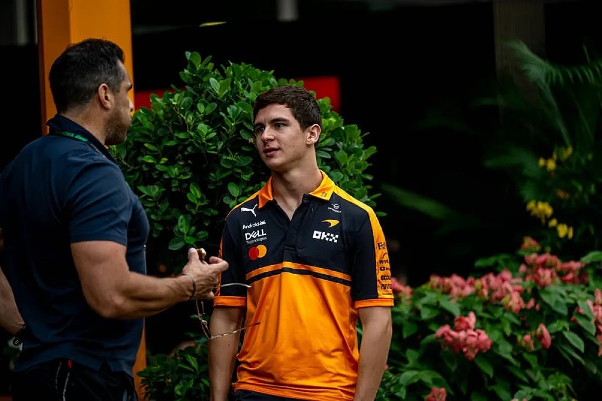 Man in McLaren orange team polo talking with a colleague in the paddock, F1 setting with green foliage behind him