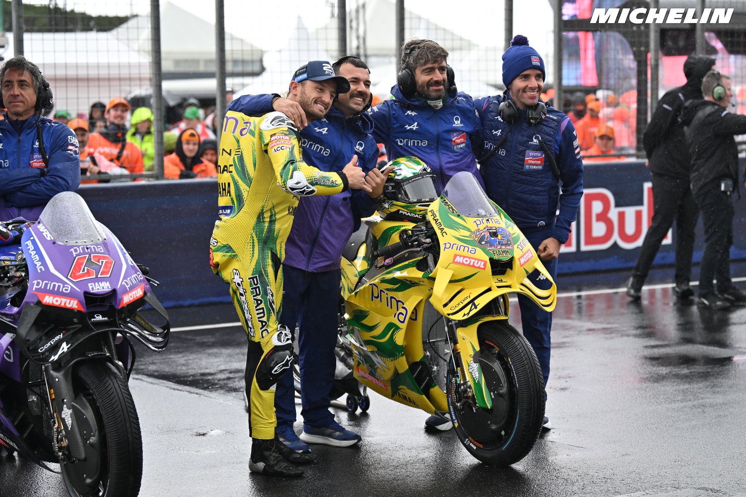Four men in team kit pose beside a yellow-and-green Prima MotoGP bike on a wet paddock, with a purple Prima bike behind them.