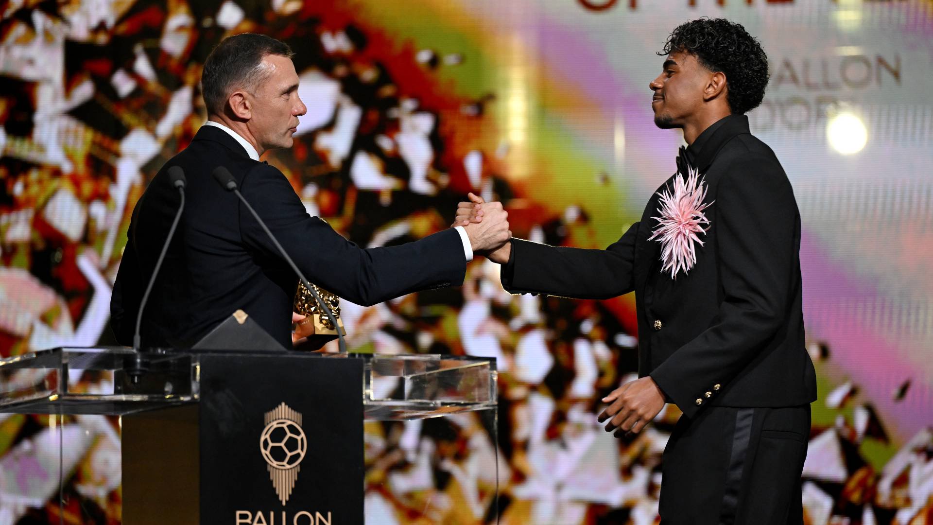Dois homens se cumprimentam no palco. Um homem caucasiano de terno escuro e camisa branca, segurando um troféu dourado, aperta a mão de um jovem negro de terno preto com uma flor rosa no peito. Ao fundo, um telão com confetes e a frase OF THE YEAR