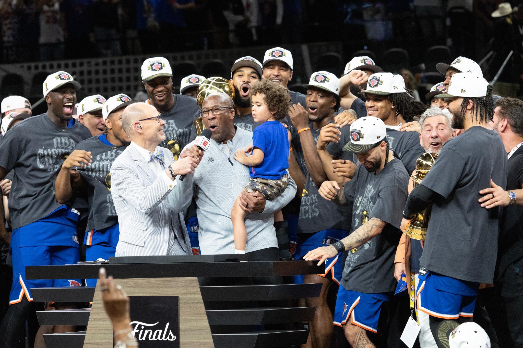New York Knicks head coach Mike Brown holds a child and takes a microphone from Ernie Johnson Jr. during the championship trophy ceremony, surrounded by celebrating players.
