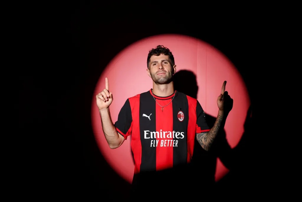 Christian Pulisic in an AC Milan jersey, pointing upwards in front of a red spotlight.