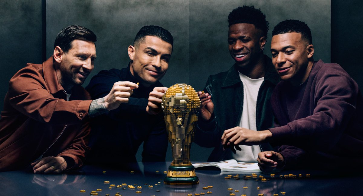A photo of footballers Lionel Messi, Cristiano Ronaldo, Vinicius Junior and Kylian Mbappe sitting around a table smiling at a LEGO model of the FIFA Football World Cup. Messi, Ronaldo and Mballe have one hand up each towards the trophy and are wearing dark tops. Vinicius Junior is wearing a black top and white t-shirt