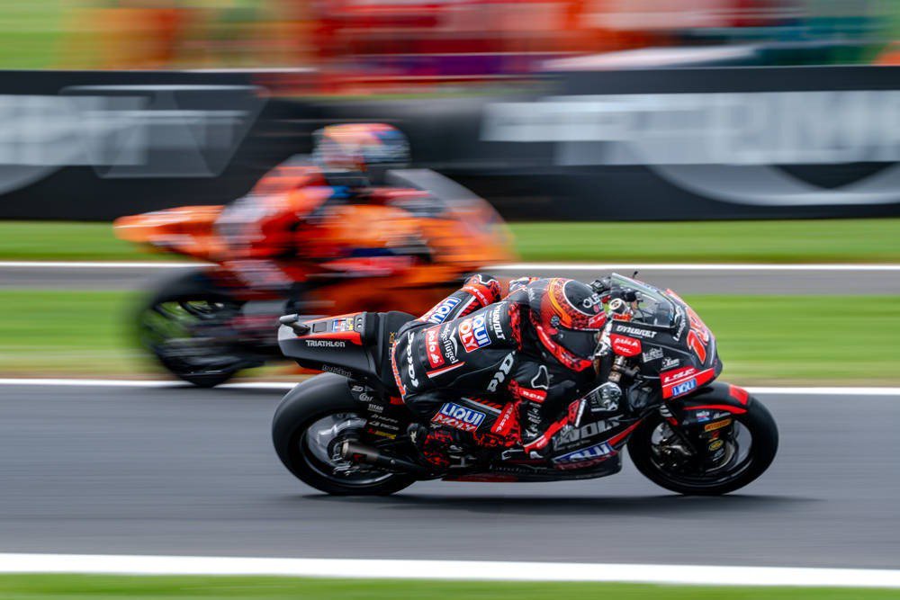Rider in black and red leathers on a black-and-red Moto2 bike leaning through a corner, with a blurred orange bike behind; Australia.