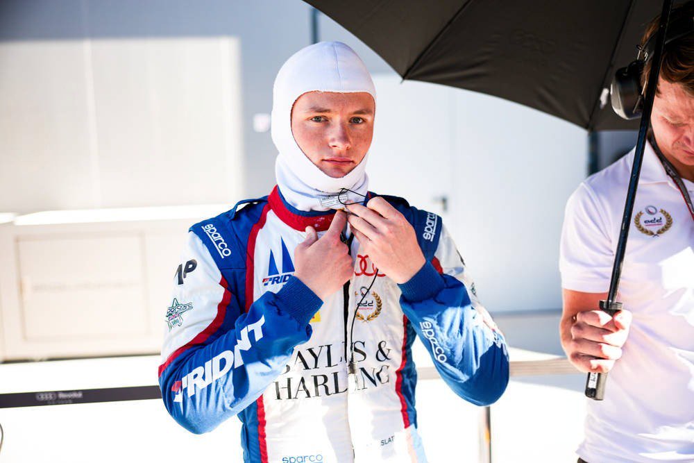 Racing driver in white balaclava and blue-red Trident suit, Bayls & Harling logos, adjusting collar in the paddock beside an umbrella-holding crew member