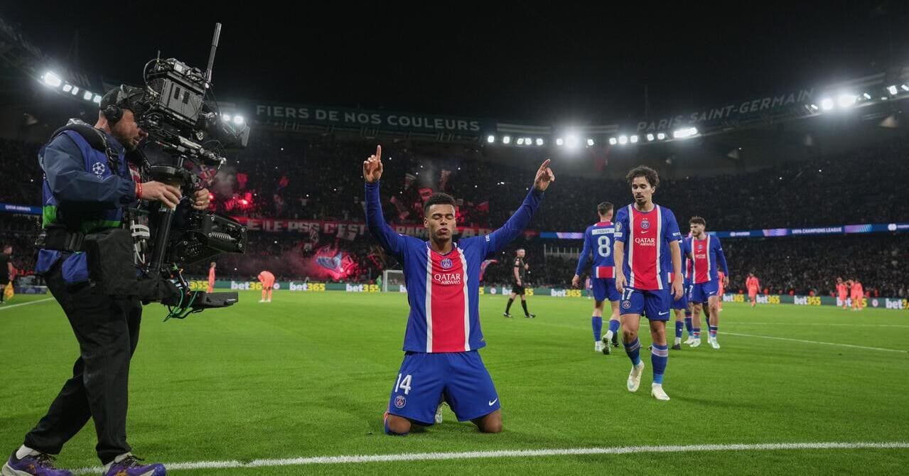 France footballer Desire Doue kneels and raising his arms to celebrate a goal while playing for Ligue 1 team Paris Saint Germain in the UEFA Champions League in the PSG home shirt of dark blue with a red stripe in the middle of two thin stripes in the middle and the words Qatar Airways in white on the chest and his number 14 in white on the shorts. Teammate Vitinha is standing to his left in the number 17 with a cameraman in a dark outfit to his left, opposition players in pink visible and fireworks coming from a crowd under lights.