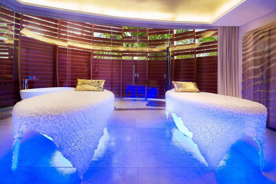 Spa de hotel com duas macas de massagem cobertas por mantas brancas, iluminação azul sob as camas e parede de madeira.