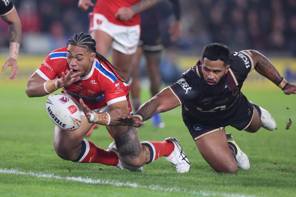 Two rugby players on green grass. One in red dives for a white ball, the other in dark jersey tackles.