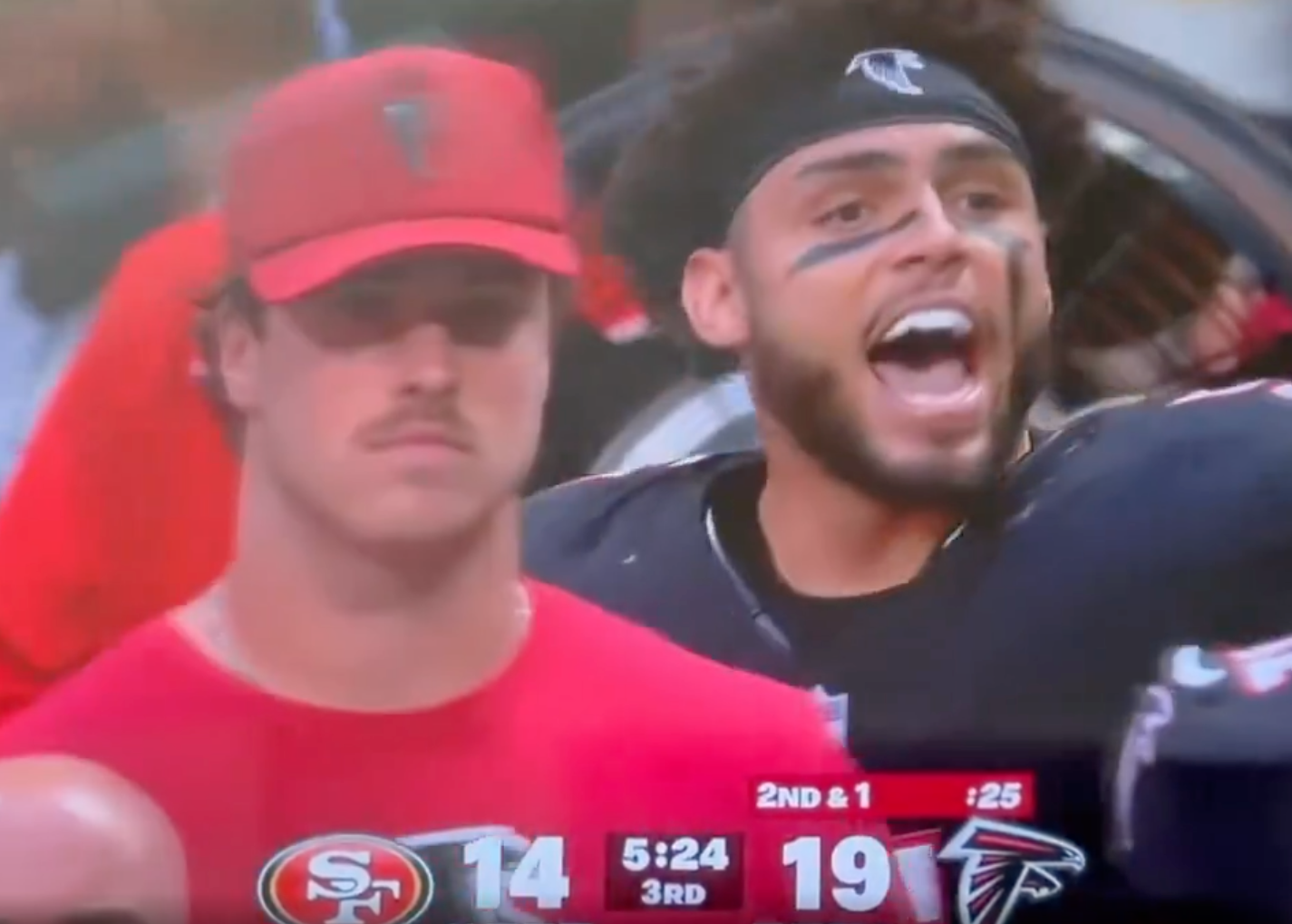 Two football players with contrasting expressions, one looking calm and the other yelling in excitement, standing on the sideline during a game.
